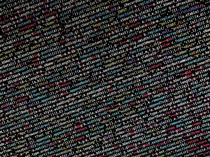 Close-up of a person's hands typing on a MacBook laptop positioned on a dark table. The screen displays a code editor with lines of programming code in various colors, including green, blue, and white