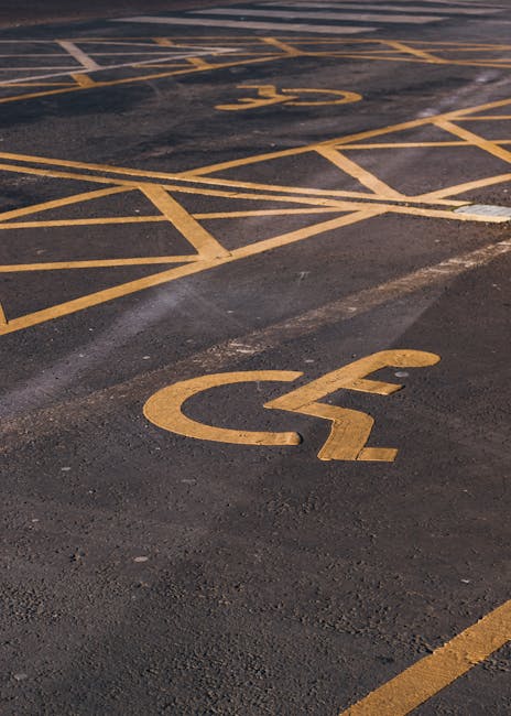 Close-up view of a section of an outdoor parking lot with yellow painted markings on dark asphalt surface, including a prominent disabled parking symbol and various diagonal lines indicating designate