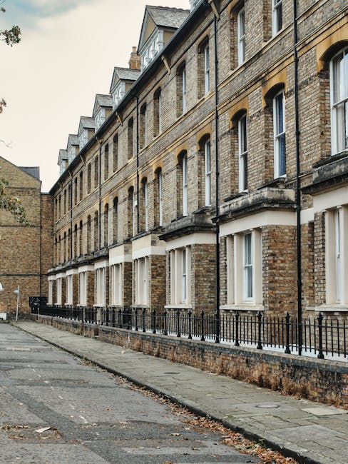 View of a narrow residential street with a row of multi-storey brick townhouses featuring large sash windows and stone window sills. The buildings have a traditional Victorian architectural style with