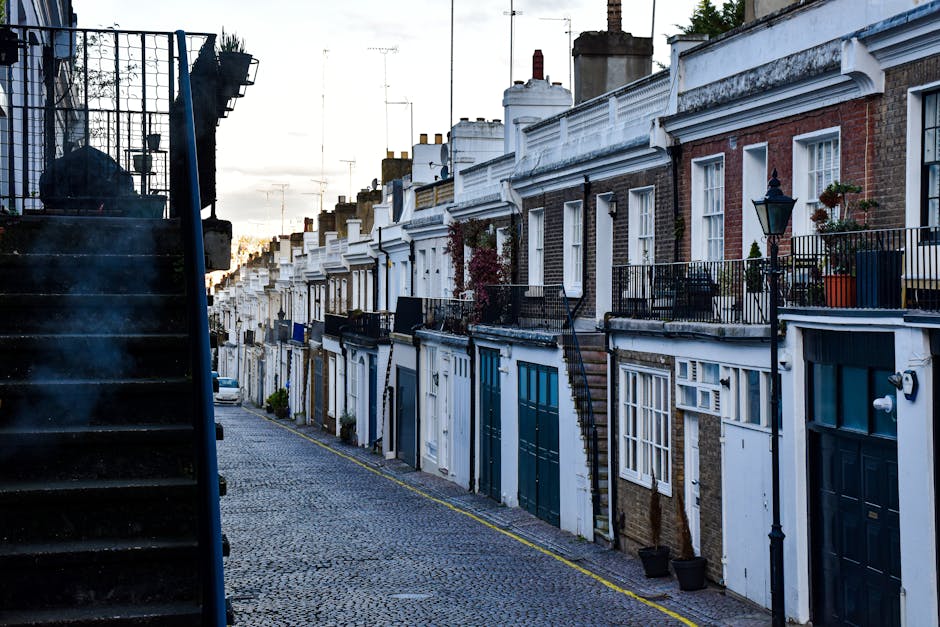 View of a narrow residential street with a row of multi-storey brick townhouses featuring large sash windows and stone window sills. The buildings have a traditional Victorian architectural style with