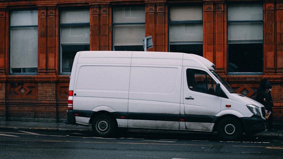 A white commercial van belonging to Man and Van Clapham Common is parked parallel along a street in front of a red brick building with large, multi-pane windows framed by ornate decorative trim. The v